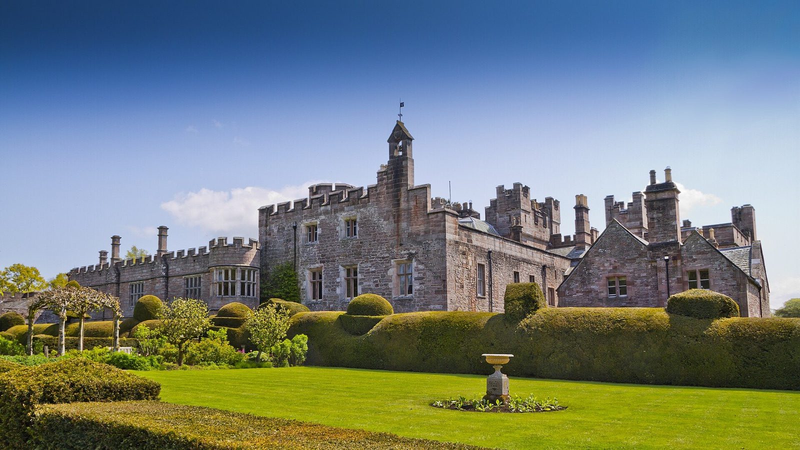 A Cumbrian country house with battlements, a bell tower and topiary hedges on a sunny day, standing in for the fictional Hollowbeck Hall